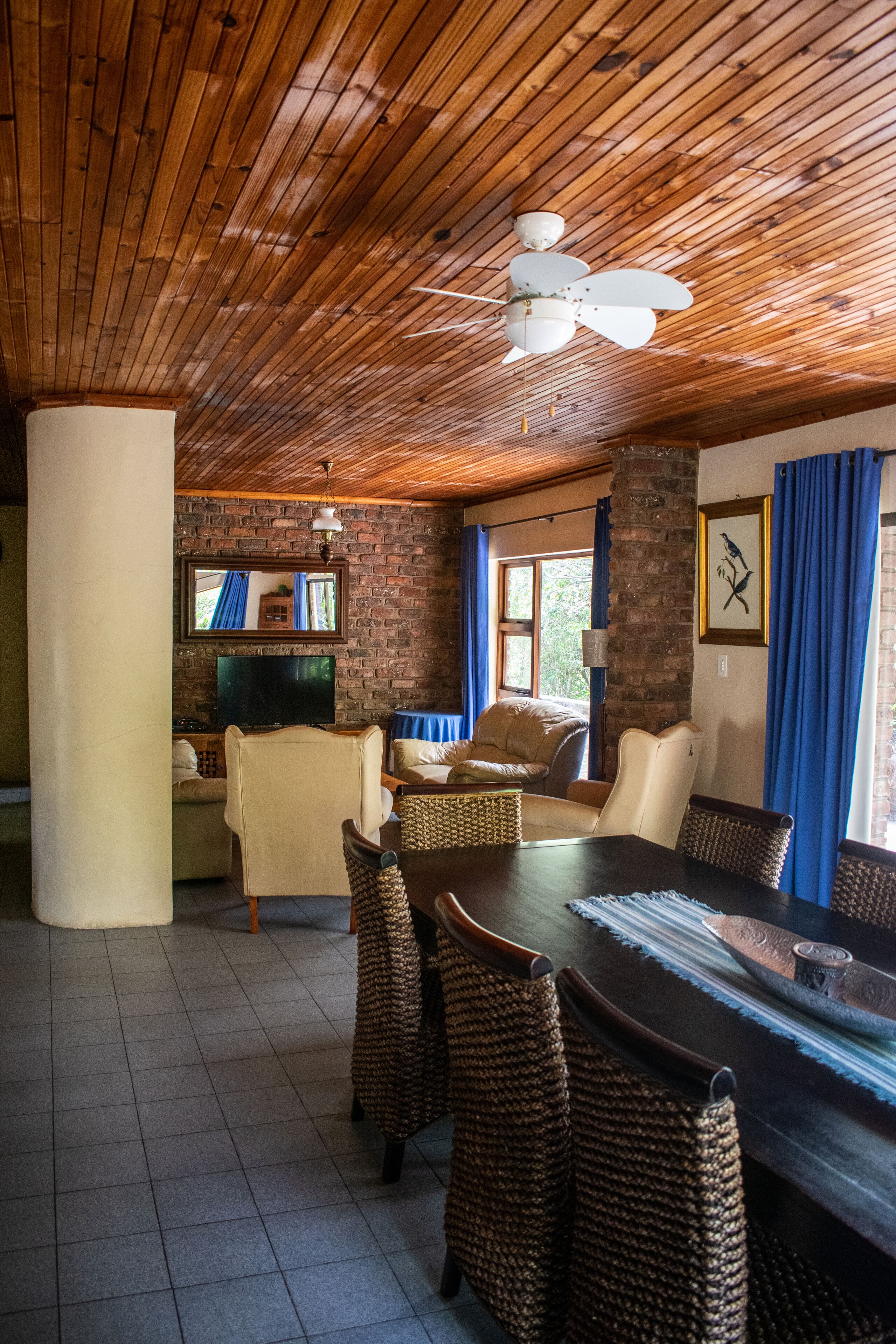 A cozy room with a wooden ceiling, a television, a dining table with wicker chairs, and a brick wall.