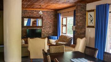 A cozy room with a wooden ceiling, a television, a dining table with wicker chairs, and a brick wall.