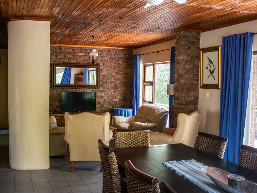 A cozy room with a wooden ceiling, a television, a dining table with wicker chairs, and a brick wall.