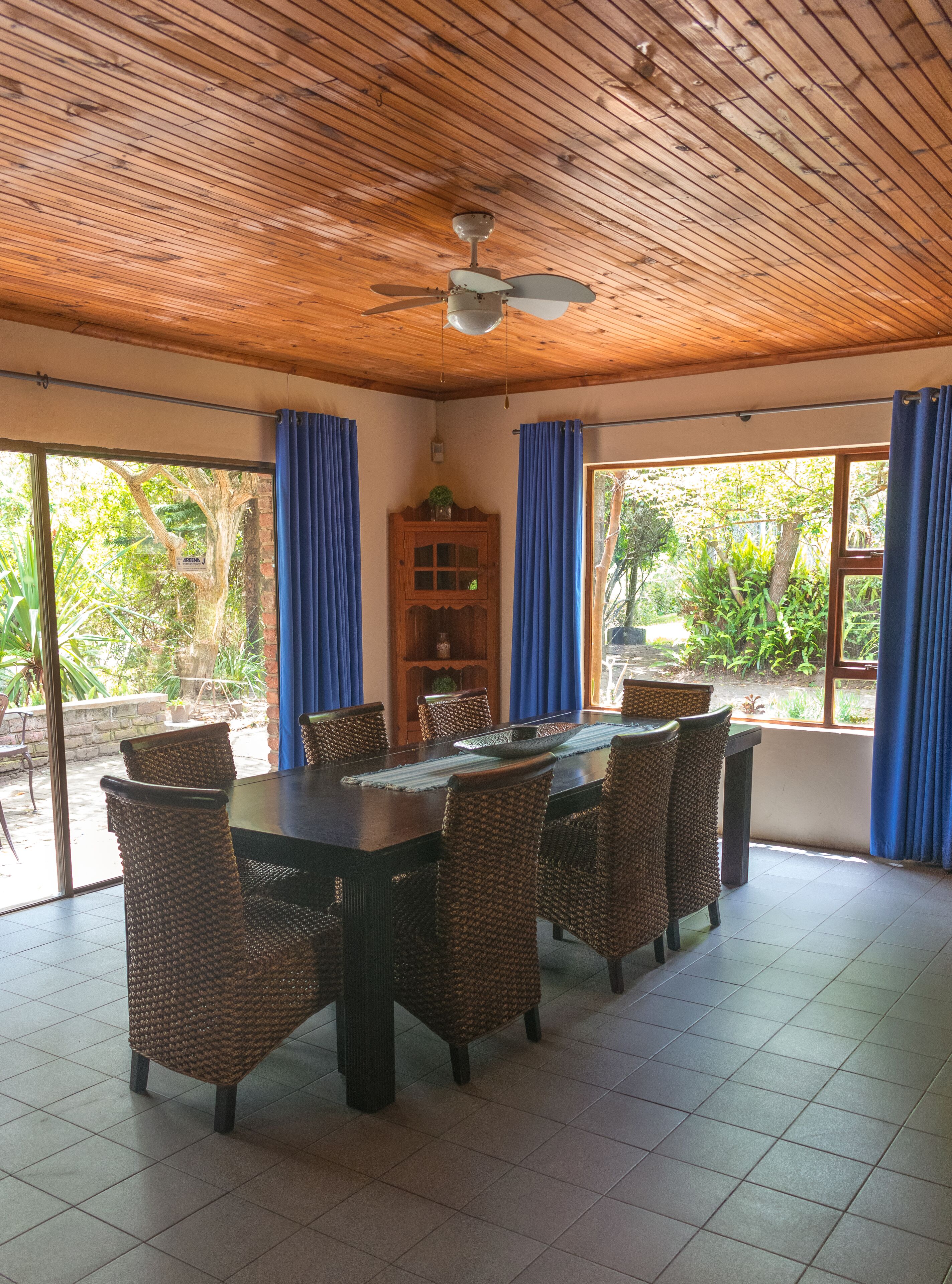 A dining area with a wooden ceiling, large windows with blue curtains, wicker chairs, and a central table.