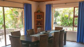 A dining area with a wooden ceiling, large windows with blue curtains, wicker chairs, and a central table.