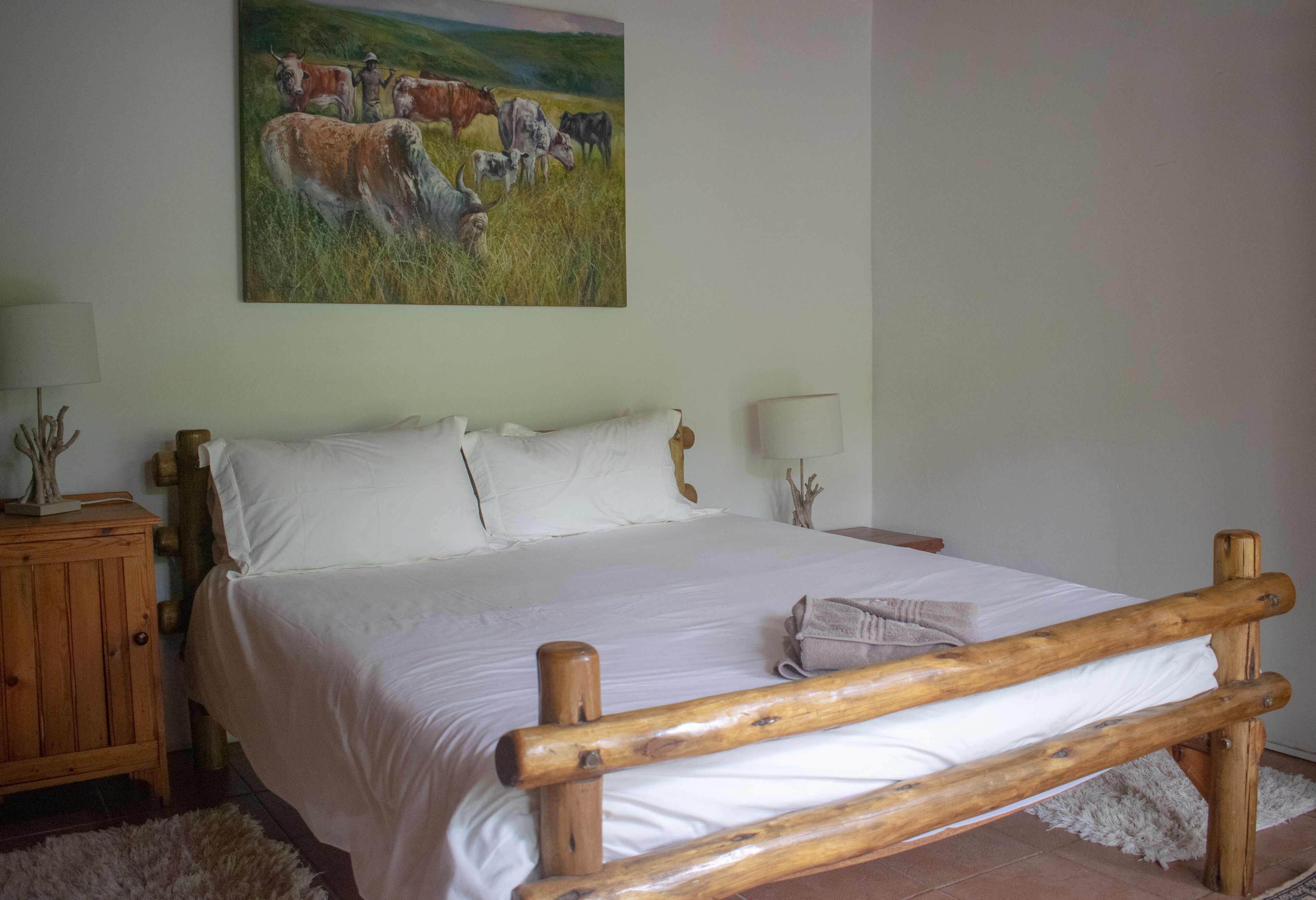 A bedroom with a wooden bed frame, white bedding, a painting of cattle on the wall, and a wooden nightstand with a lamp.