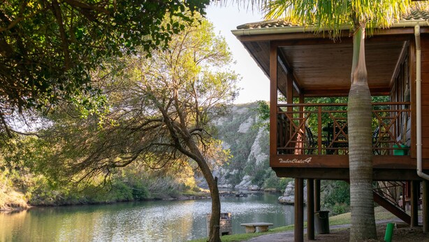 Une structure en bois dotée d’un balcon donnant sur un lac paisible, entourée d’une végétation luxuriante et d’une falaise rocheuse.