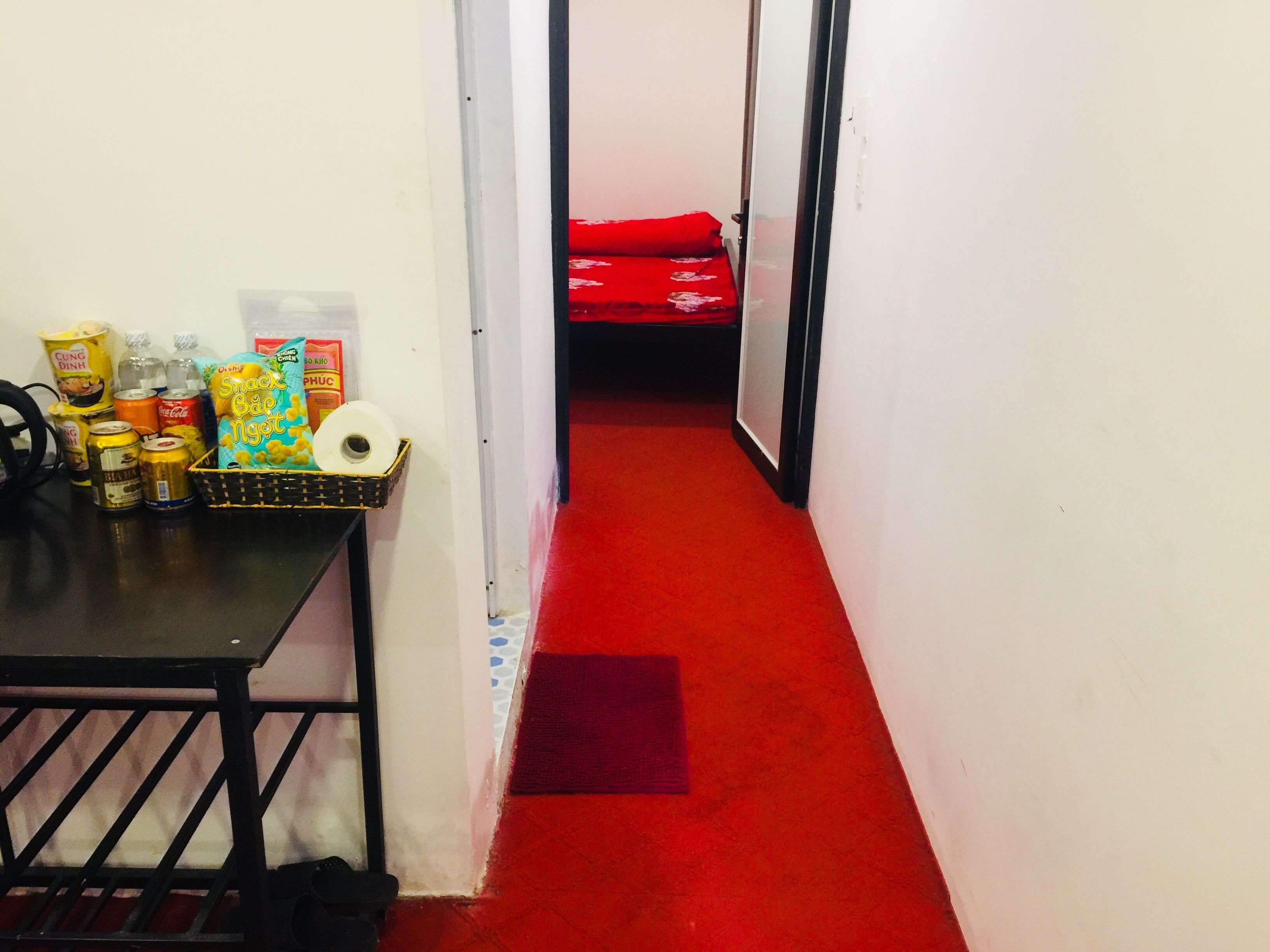 A narrow hallway with a red carpet, a small table with snacks and drinks, and a bed with red bedding in the background.