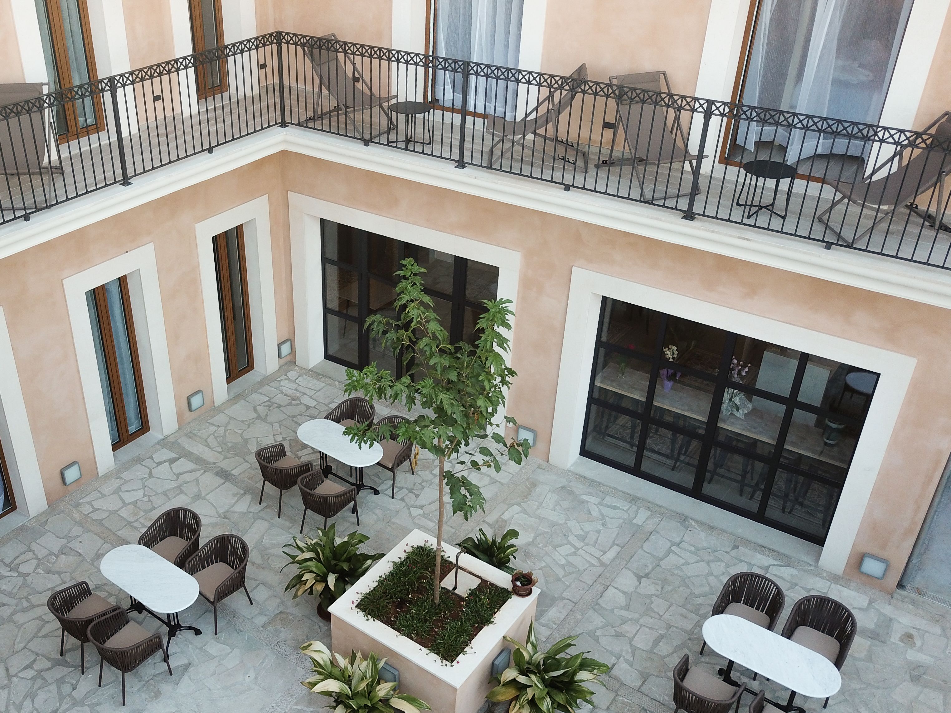 Un patio con mesas al aire libre, una jardinera central con un árbol y un balcón con barandilla.