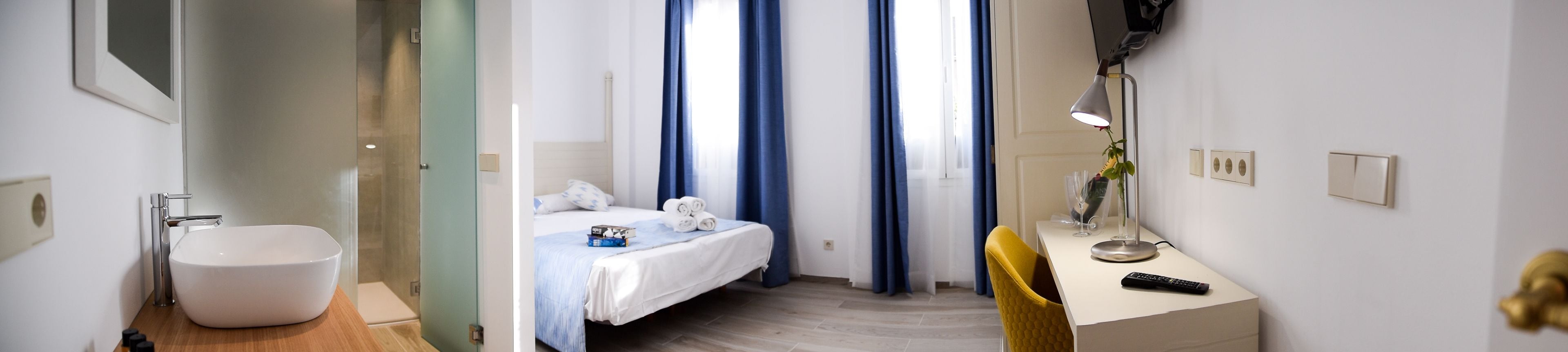 Een hotelkamer met een wit wastafel, een bed met blauw beddengoed, een bureau met een lamp en een stoel.