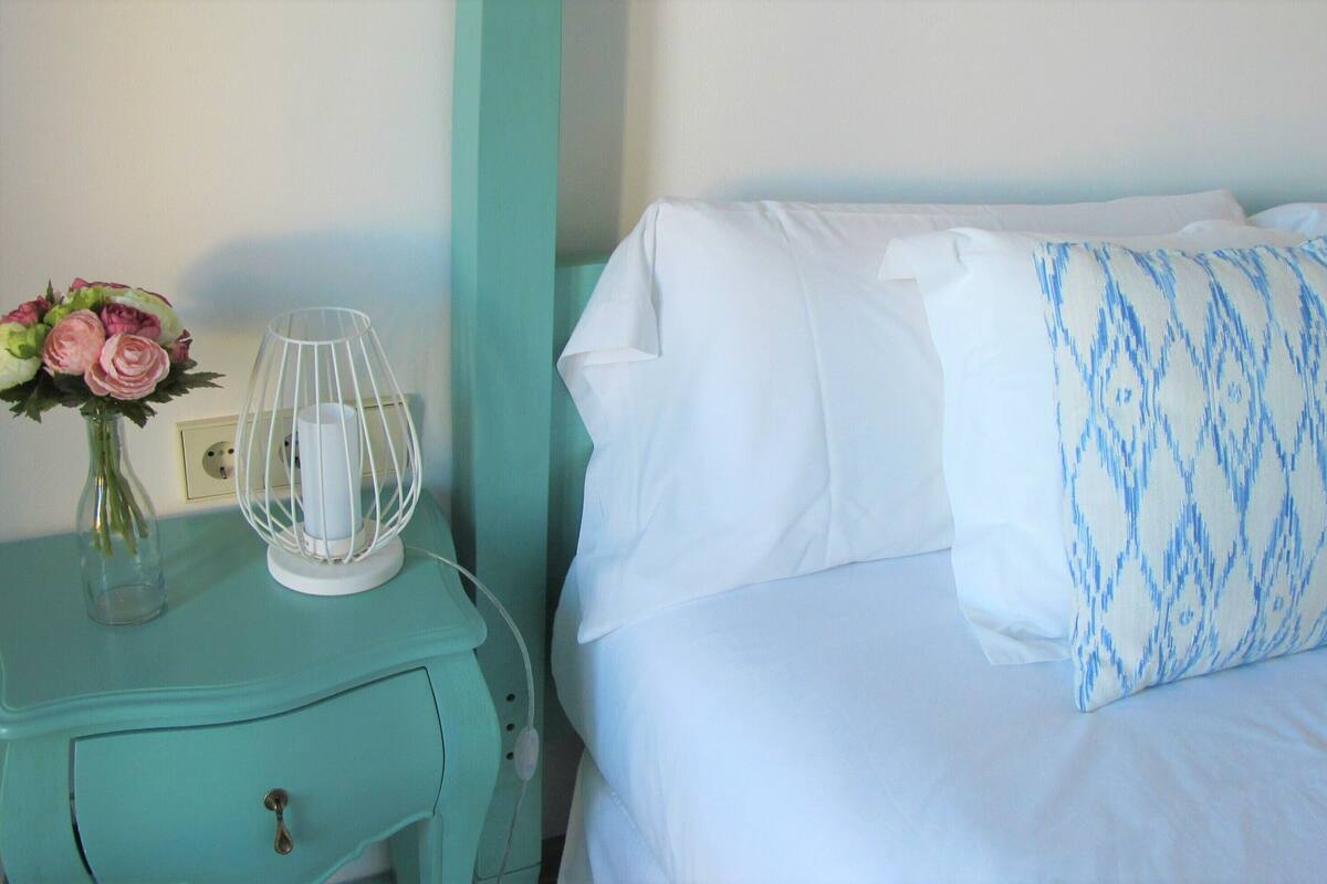 A bed with white bedding and a patterned pillow, a teal nightstand with a vase of flowers and a lamp, and a wall socket.