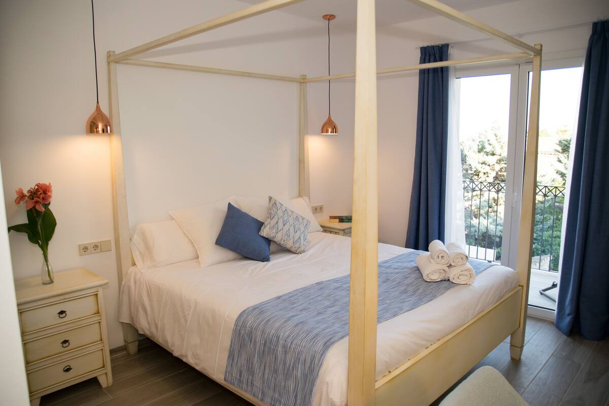 A four-poster bed with a white and blue bedspread, a bedside table with a vase of flowers, and a view of the outdoors through a window with blue curtains.