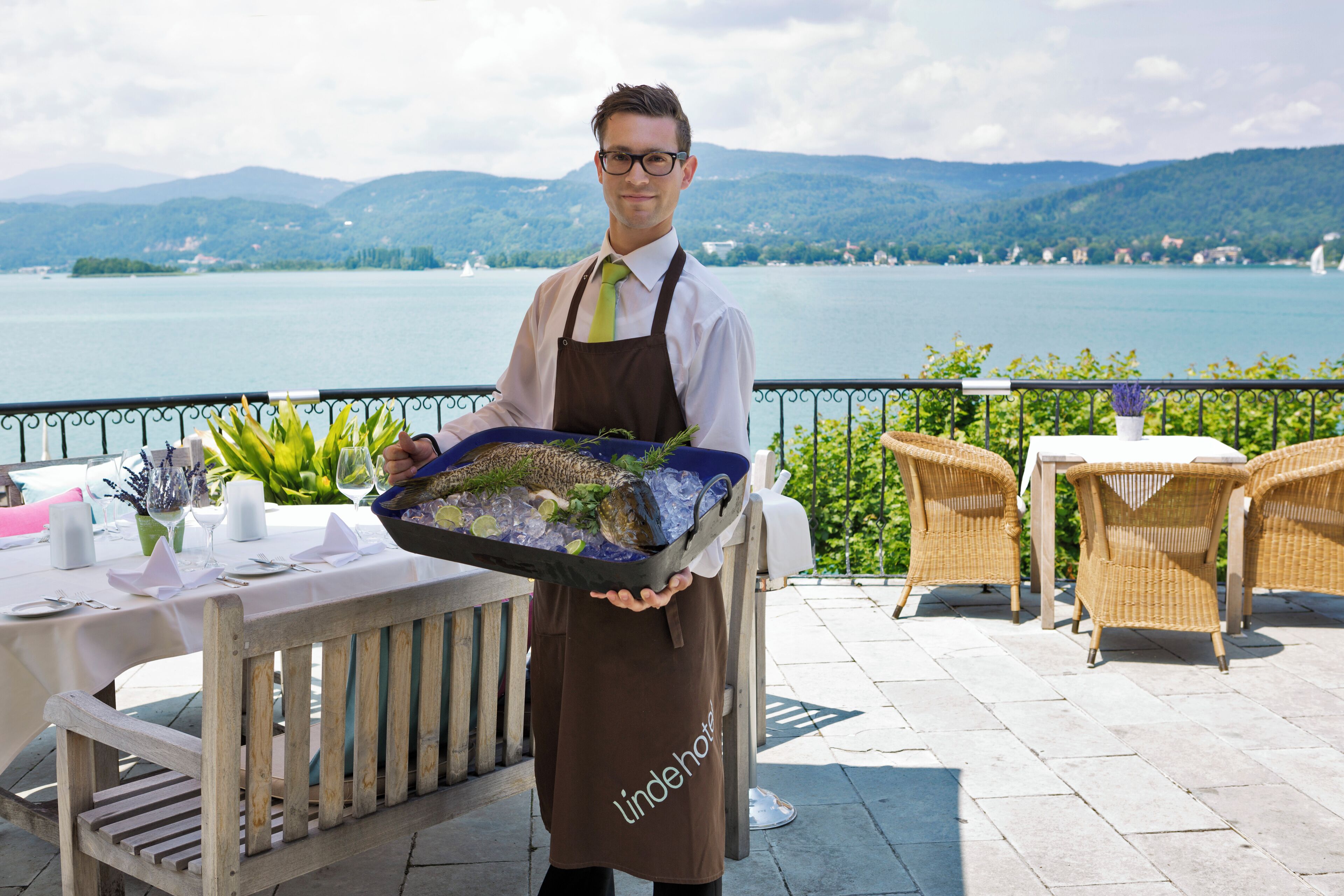 A waiter holding a tray with food and drinks, standing on a terrace with tables and chairs, overlooking a lake and mountains.
