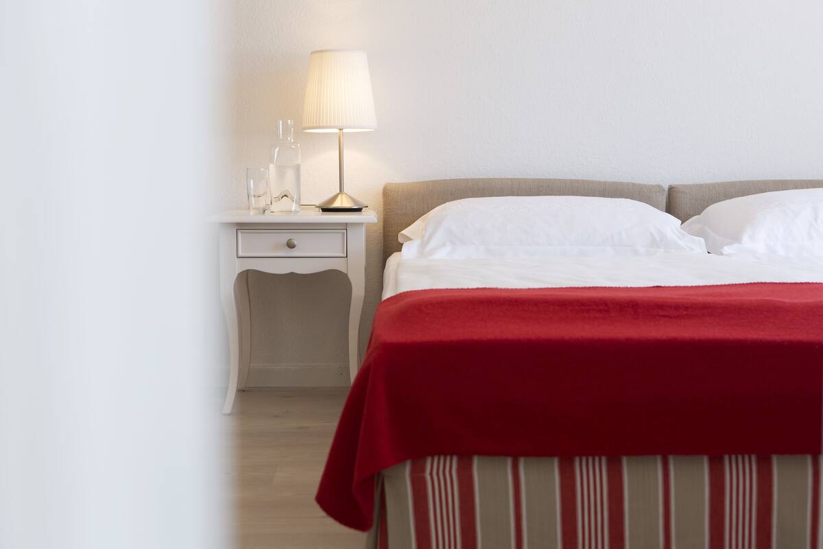 Ein ordentlich gemachtes Bett mit weißen Laken und einer roten Decke, ein Nachttisch mit Lampe und Gläsern sowie eine schlichte Wand.