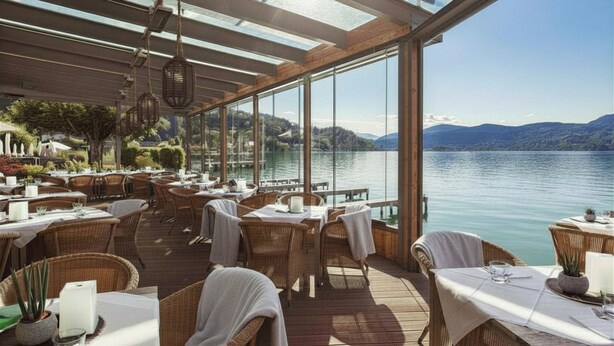An outdoor dining area with wicker furniture and tables set for dining, overlooking a calm lake and distant mountains. - Hotel Linde (Maria Woerth)