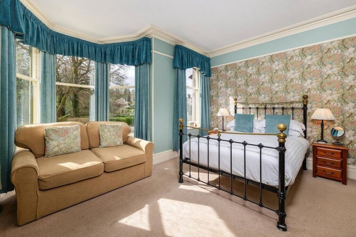 Un dormitorio con una cama, una mesita de noche de madera, un sofá beige y una ventana con cortinas.