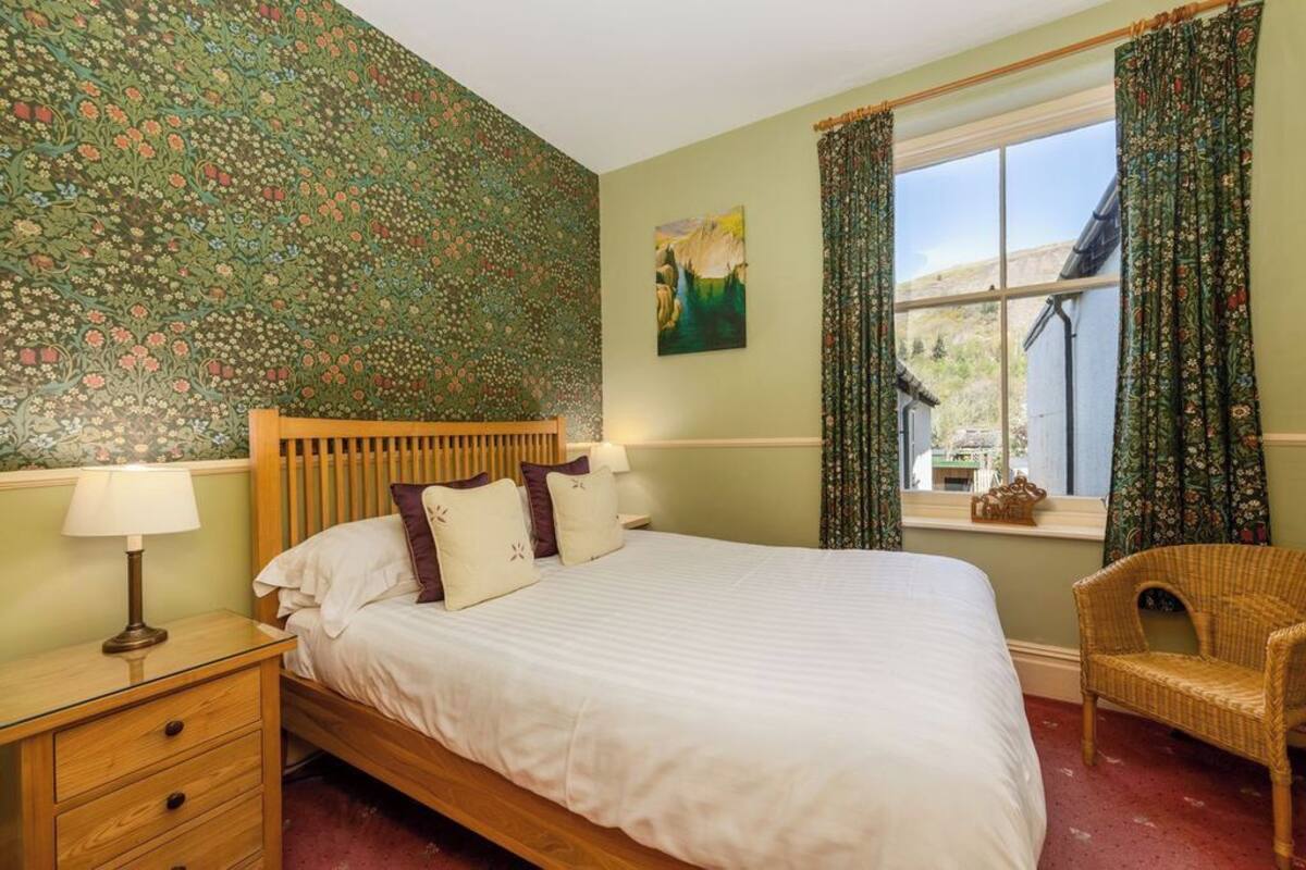 Un dormitorio con papel tapiz floral, una cama de madera, una mesita de noche, una silla de mimbre y una ventana con cortinas.