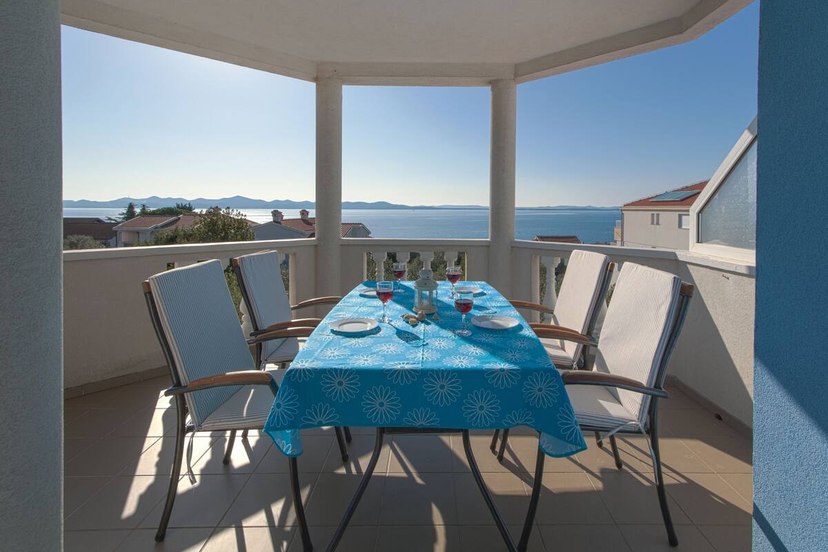 Un balcon avec une table dressée pour deux personnes, offrant une vue sur une étendue d’eau et des montagnes lointaines.