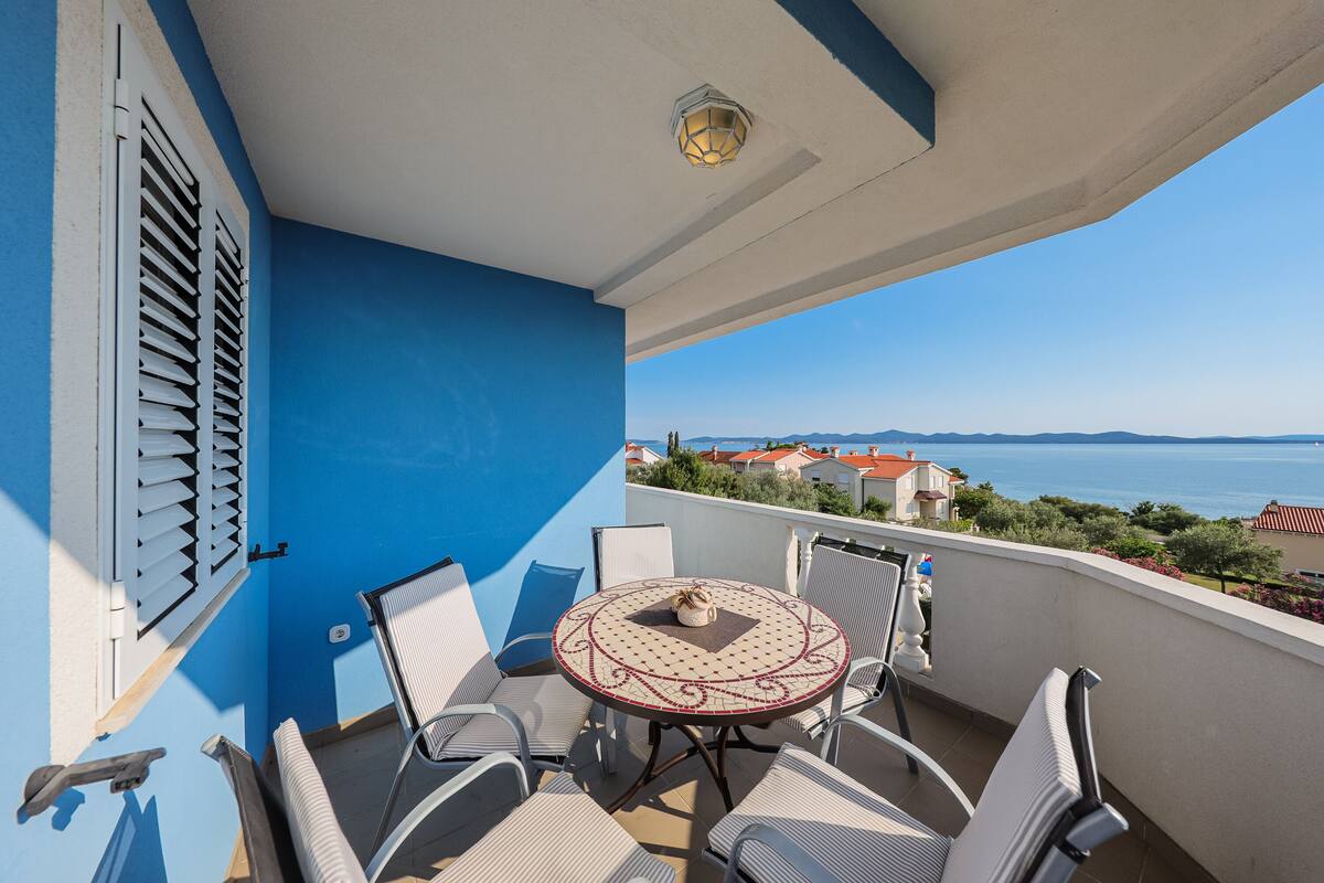 Un balcon avec une table ronde et des chaises, une vue sur la mer et, au loin, un bâtiment aux toits rouges.