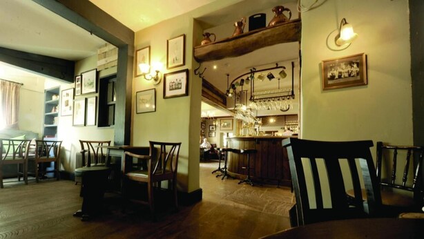 A cozy dining area with wooden furniture, a bar in the background, and framed pictures on the walls. - The Dog at Peover (Knutsford)