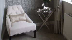 A room with a patterned wallpaper, a round mirror, a white armchair, a small table with a vase of flowers, and a window with curtains. - The Dog at Peover (Knutsford)
