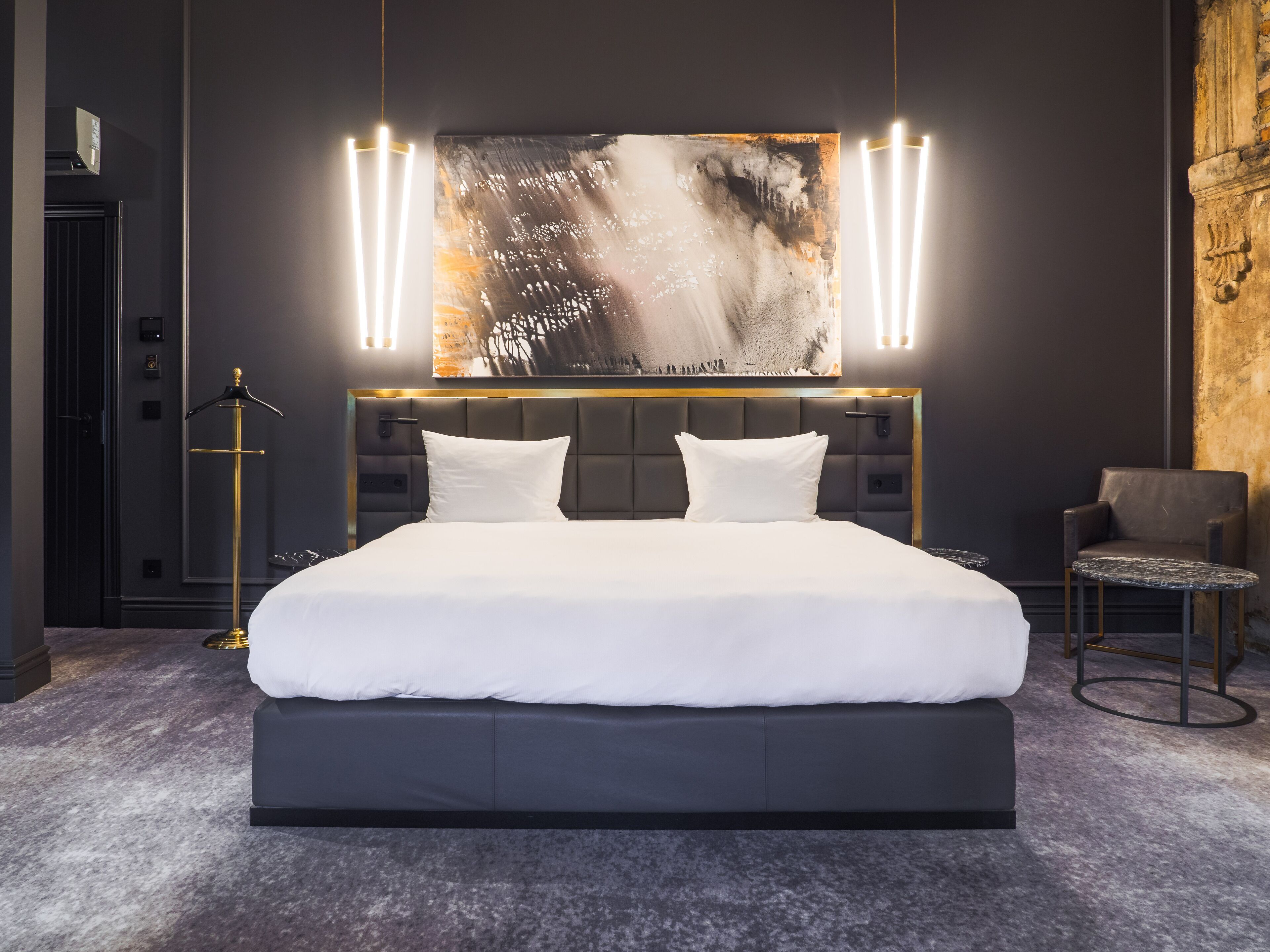 Un dormitorio moderno con una cama grande, un cabecero con iluminación integrada, un cuadro abstracto de gran tamaño sobre la cama y una mesita redonda con una silla.