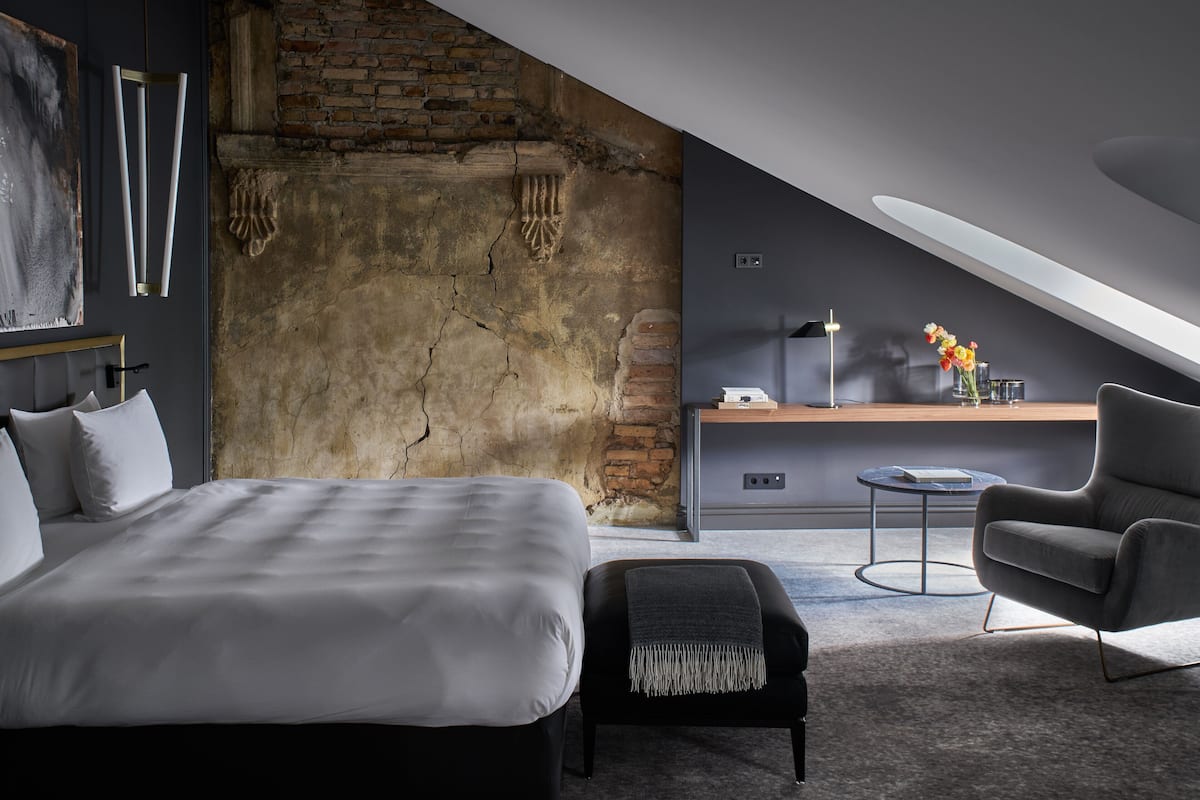 A modern bedroom with a large bed, a small bench, a chair, a desk, and a view of a brick wall and ceiling.