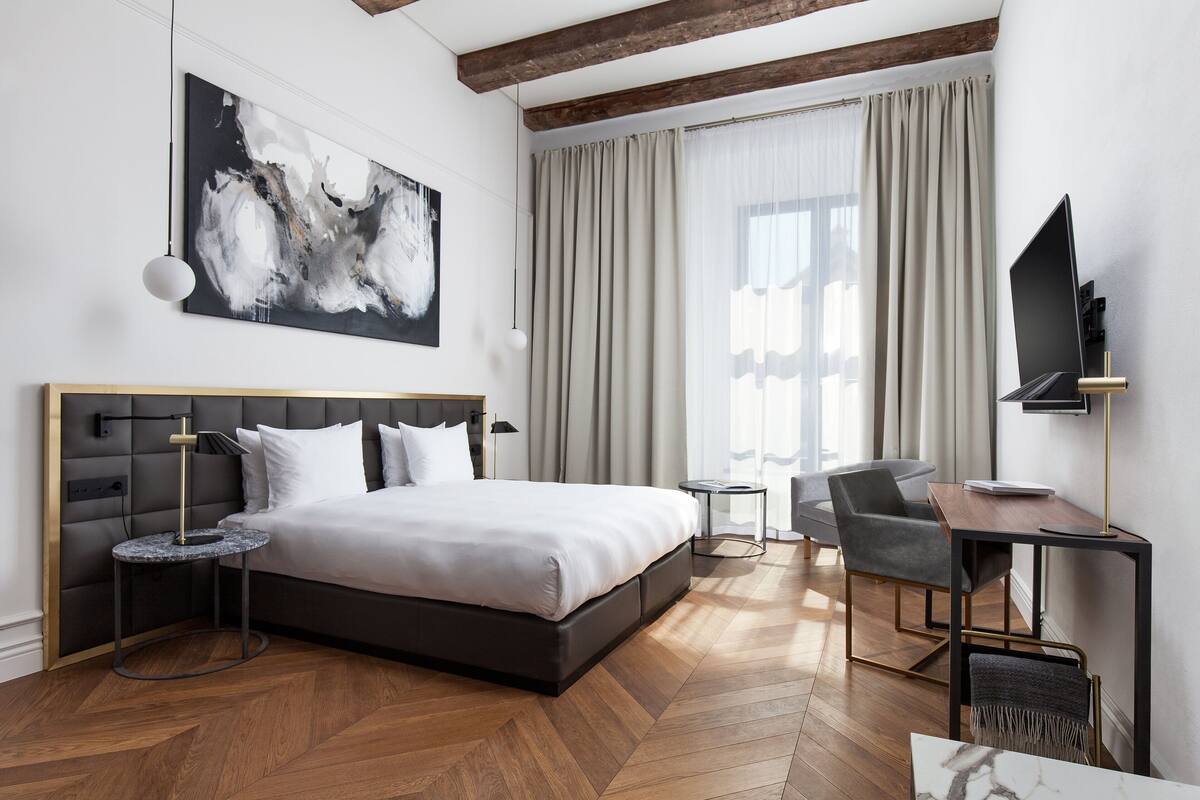 A modern hotel room with a large bed, a desk, a chair, a TV, and a painting on the wall.