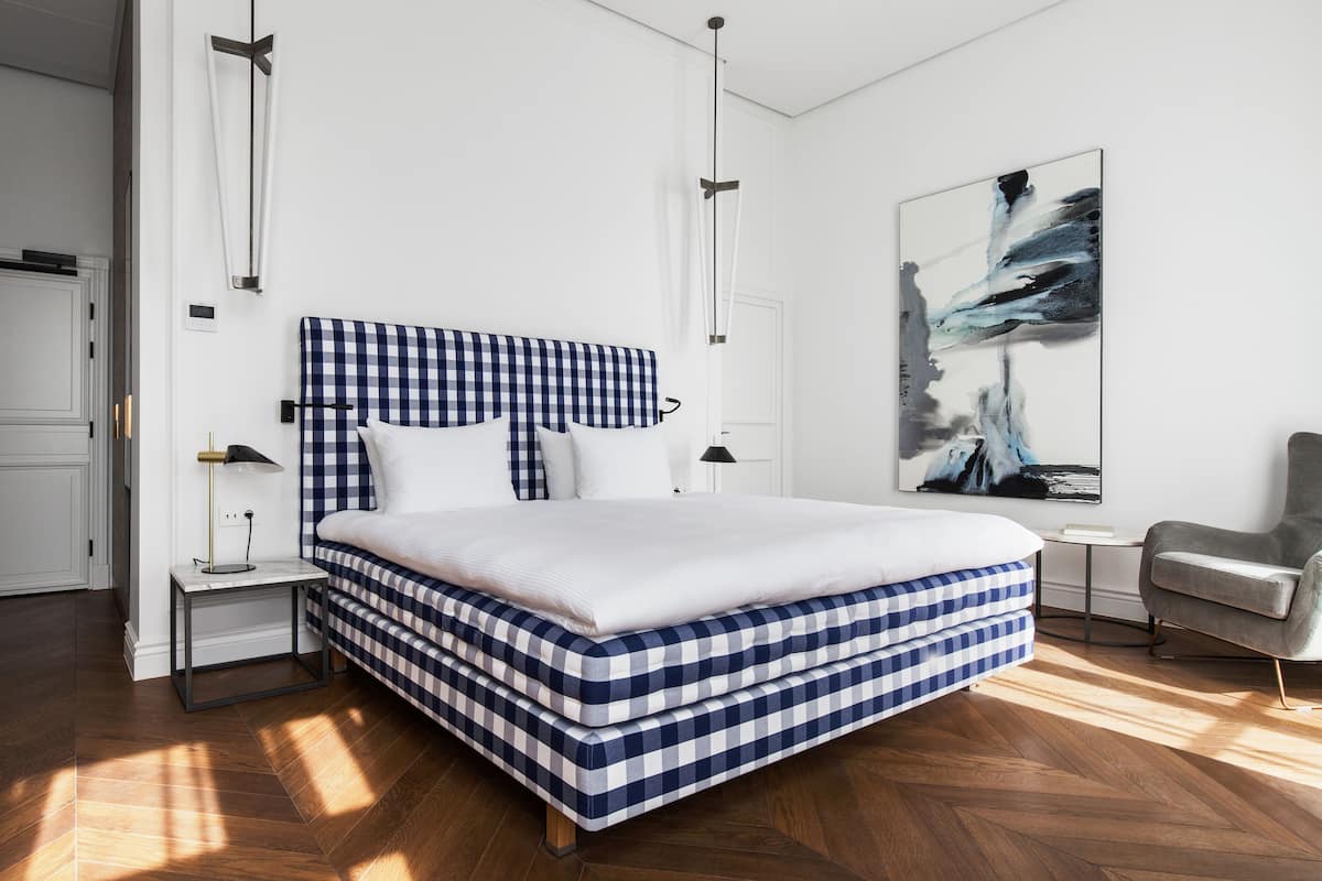 A modern bedroom with a checkered bedspread, a large abstract painting on the wall, and a grey armchair.