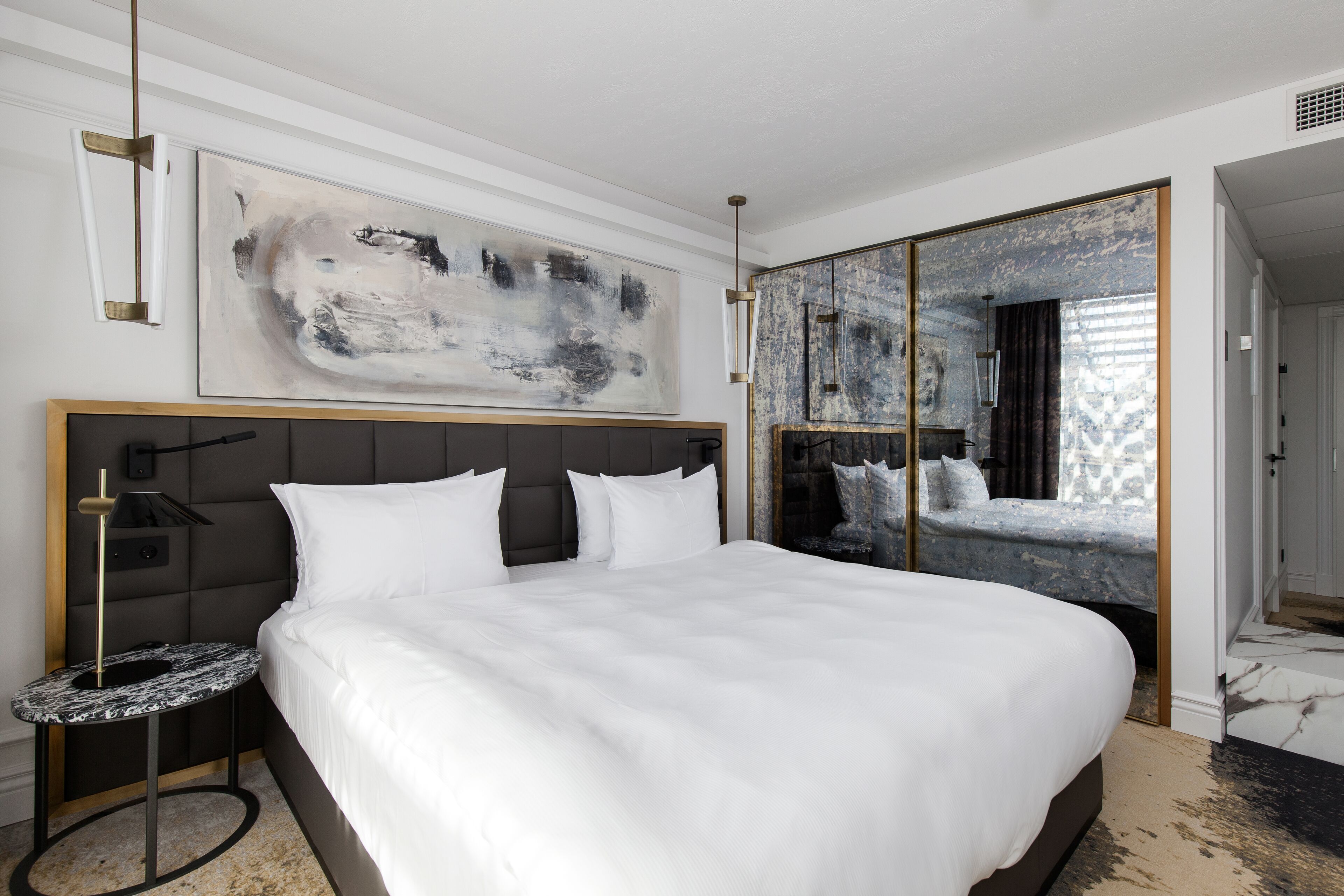 Et moderne hotelværelse med en stor seng, et sengebord, et stort abstrakt maleri og et spejlskab.