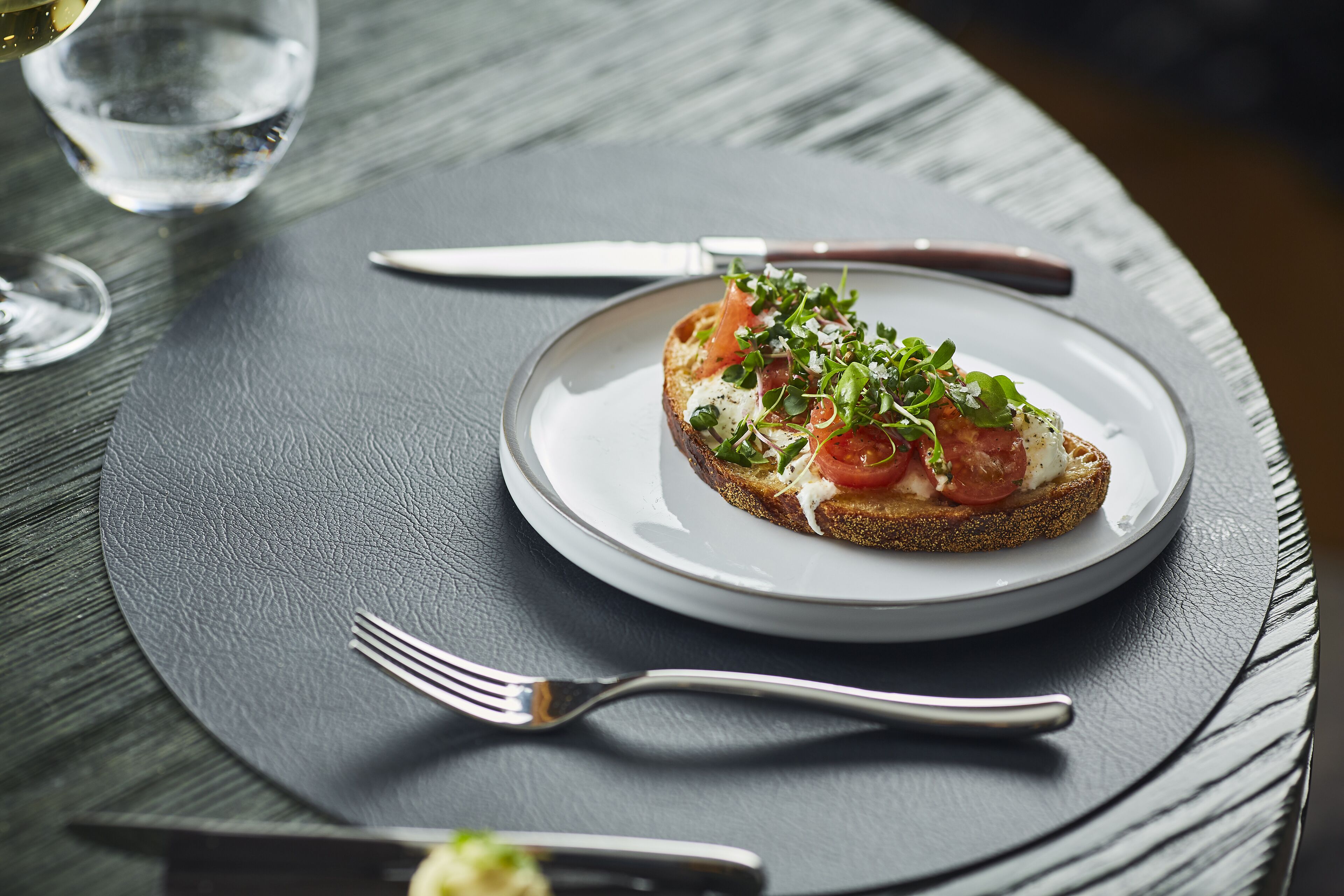 A plate with a slice of bread topped with greens and tomatoes, accompanied by a fork and knife on a dark table.