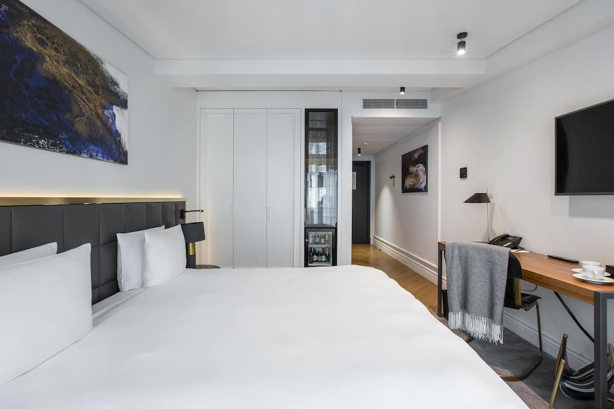 A modern hotel room with a large bed, a desk with a telephone and a cup, a TV mounted on the wall, and a painting above the bed.