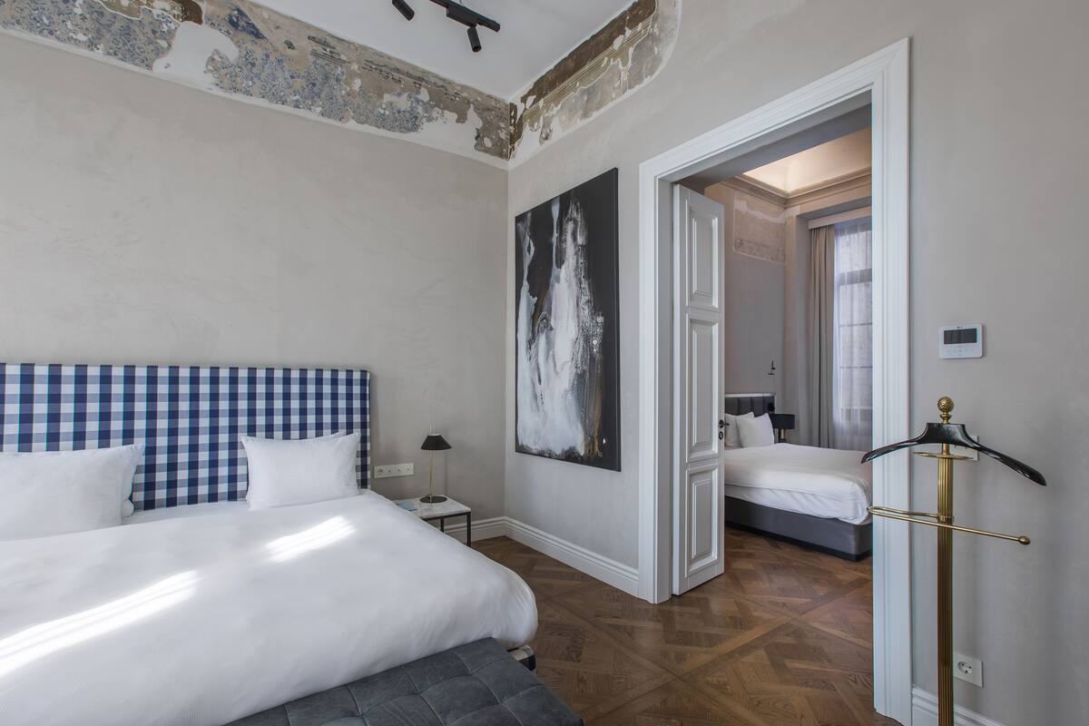 A bedroom with a checkered headboard, a large painting, a coat rack, and a view into another room.
