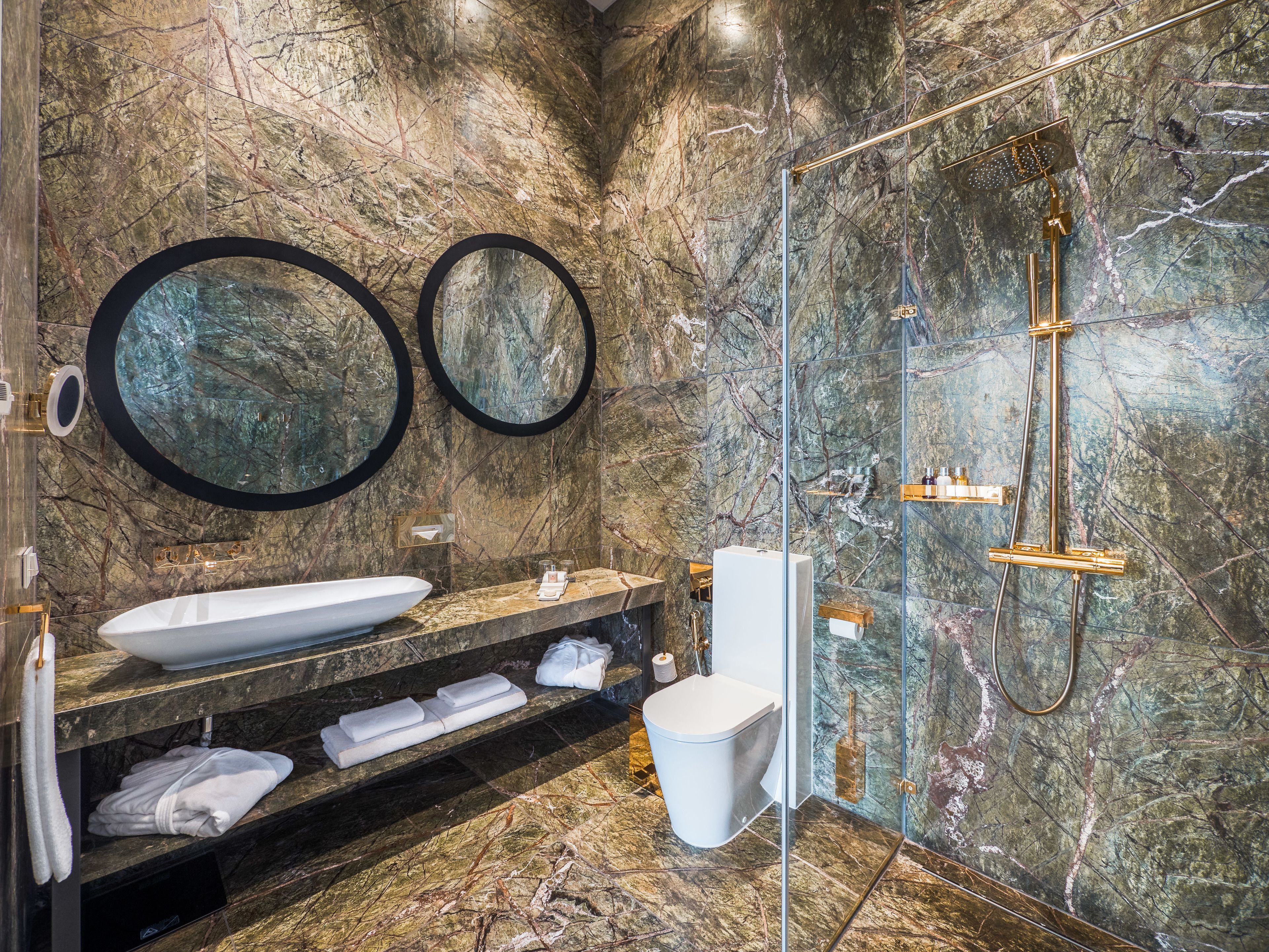 A modern bathroom with a marble wall, a large sink, two round mirrors, a glass-enclosed shower, and neatly folded towels.