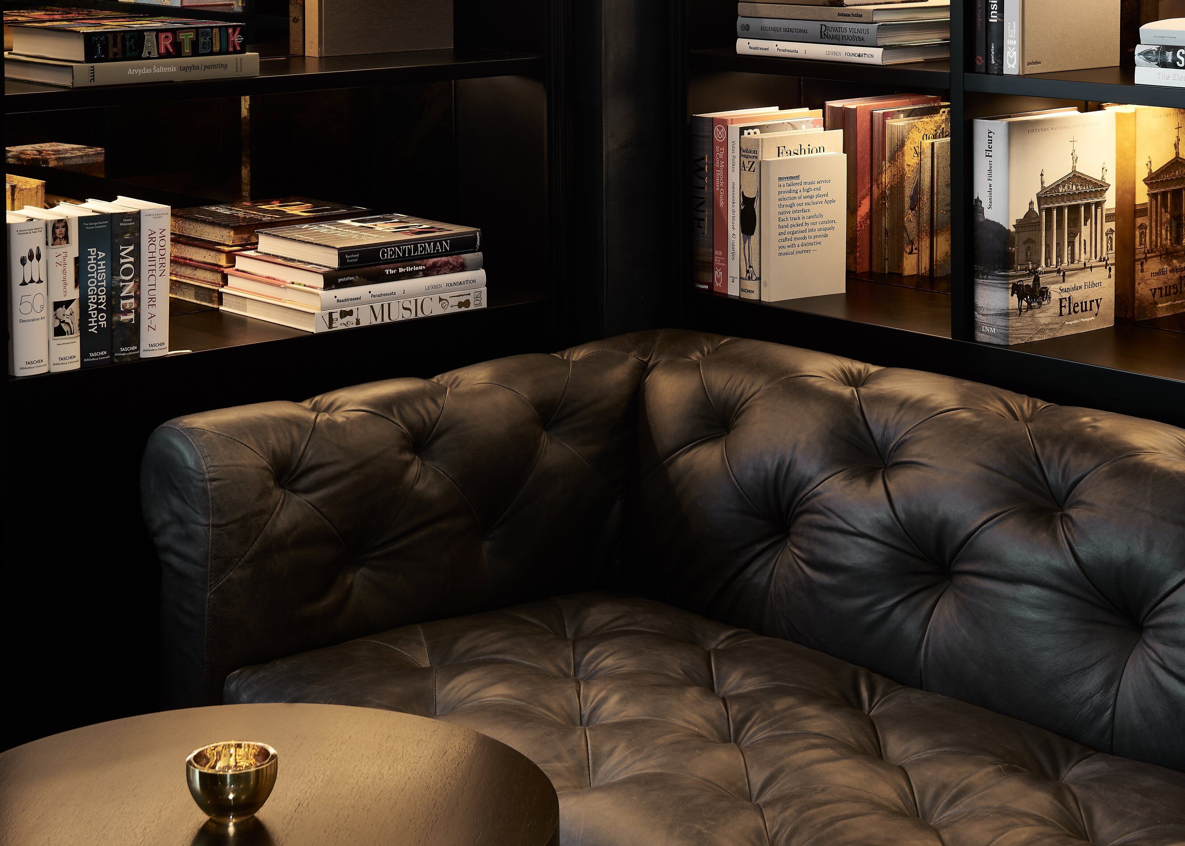 Ein Ledersessel steht in einer Ecke, mit einem kleinen Tisch und einer Schale darauf. Im Hintergrund sind Bücherregale mit verschiedenen Büchern zu sehen.