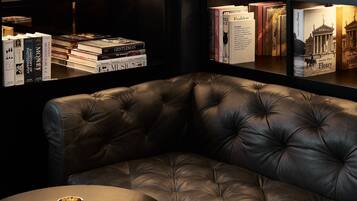 Ein Ledersessel steht in einer Ecke, mit einem kleinen Tisch und einer Schale darauf. Im Hintergrund sind Bücherregale mit verschiedenen Büchern zu sehen.