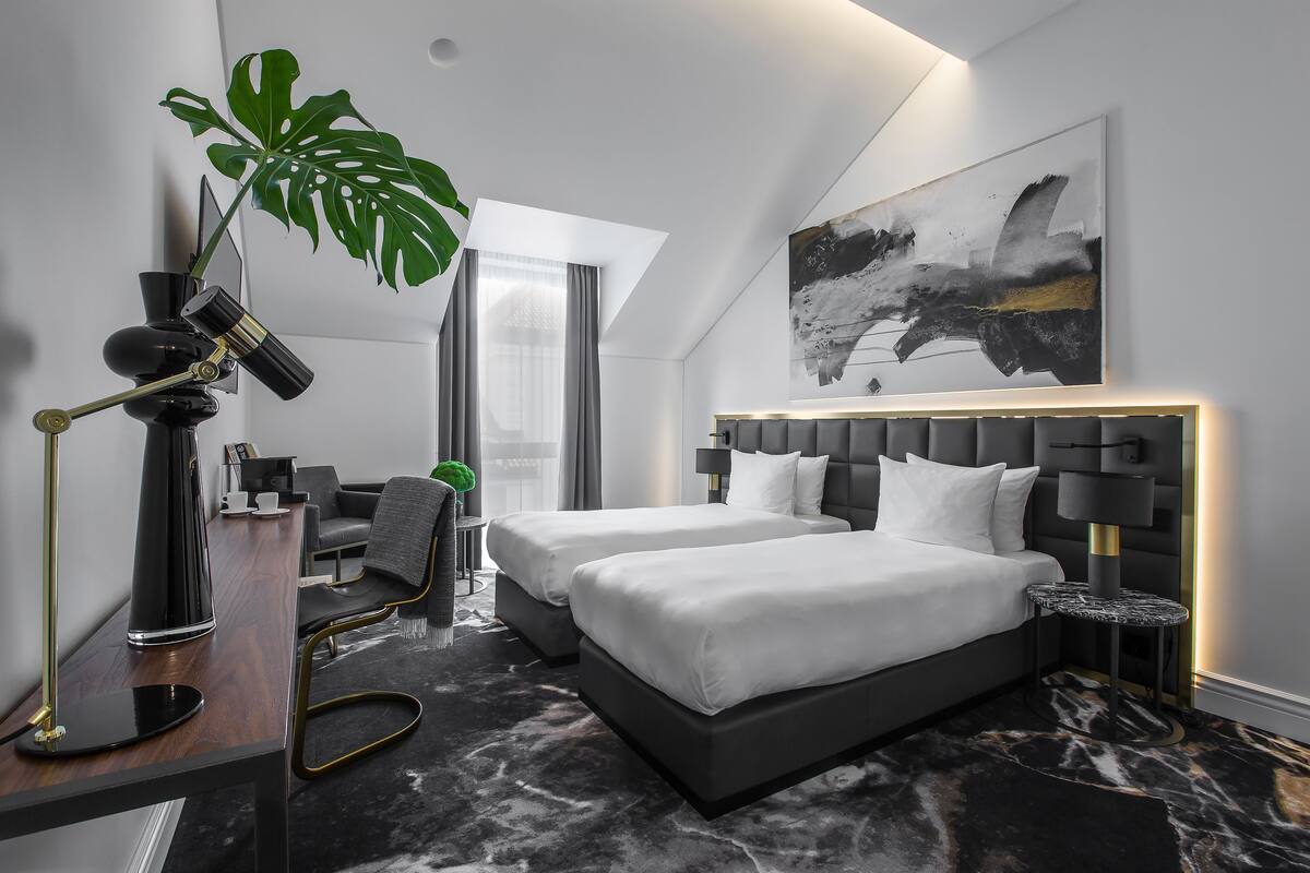 A modern hotel room with a large bed, bedside tables, a desk, a chair, a potted plant, and a large abstract painting on the wall.