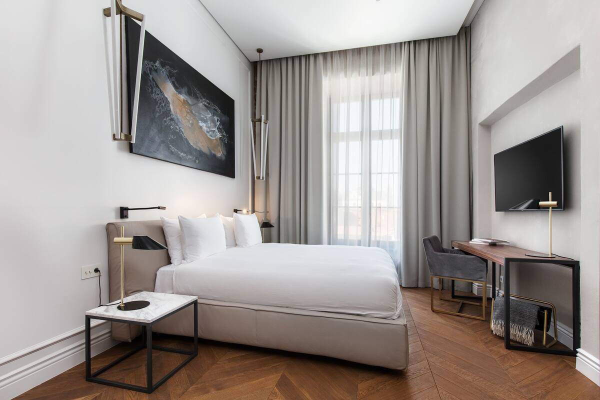 A modern hotel room with a large bed, a desk, a TV, and a painting on the wall.