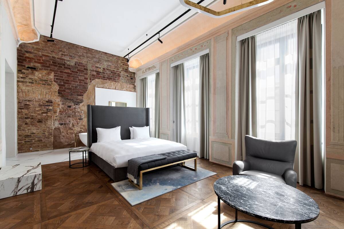 A modern hotel room with a large bed, a bench, a chair, and a marble table.
