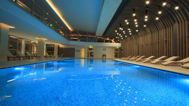 Una piscina interior con suelo de mosaico azul, sillas de descanso y un diseño arquitectónico moderno con techo curvo e iluminación empotrada.