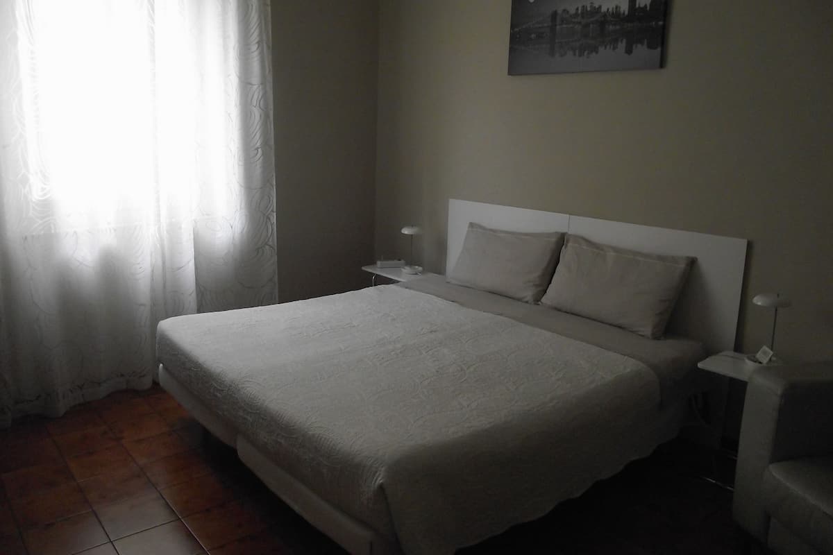 Une chambre avec un grand lit, une table de chevet, une lampe et un tableau représentant un paysage urbain accroché au mur.