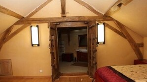 A bedroom with a sloped ceiling, wooden beams, a bed with a red quilt, and a door leading to a bathroom. - Château de Belle Poule  (Les Ponts-de-Ce)