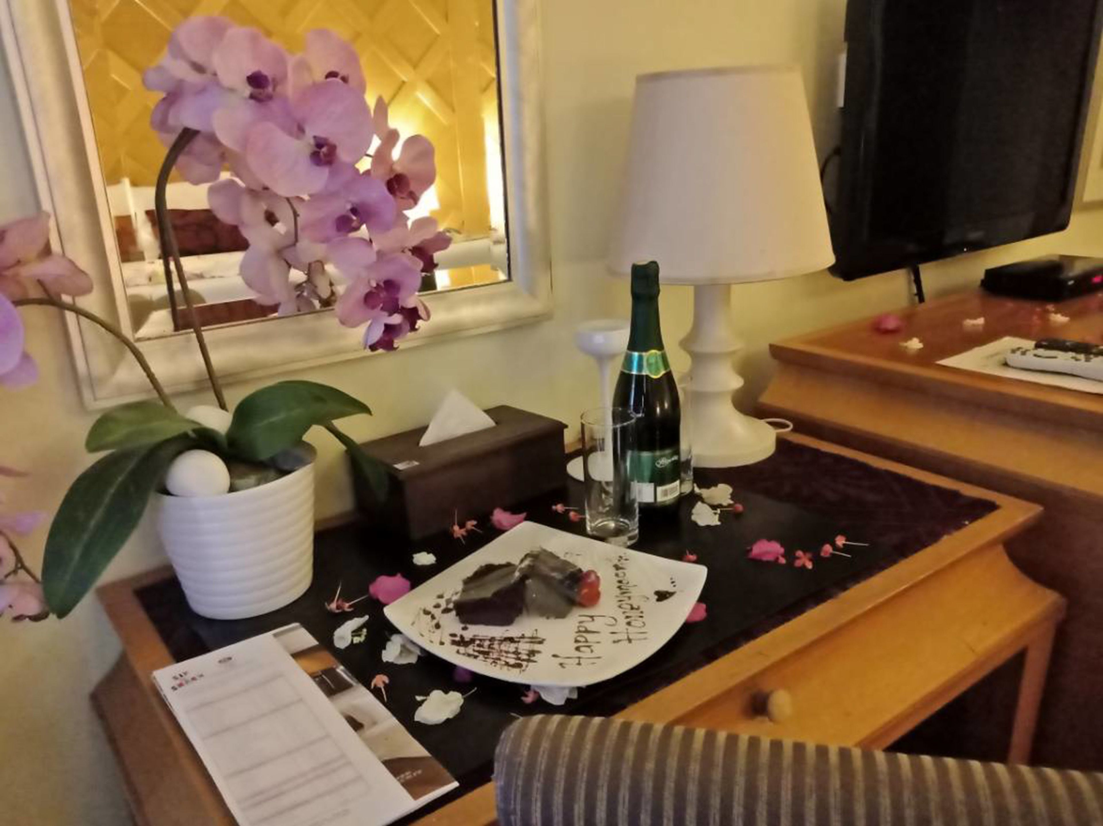 Habitación de hotel con una cama, una mesita de noche con una botella de champán, una caja de pañuelos, un plato con un postre de chocolate y una orquídea en maceta.