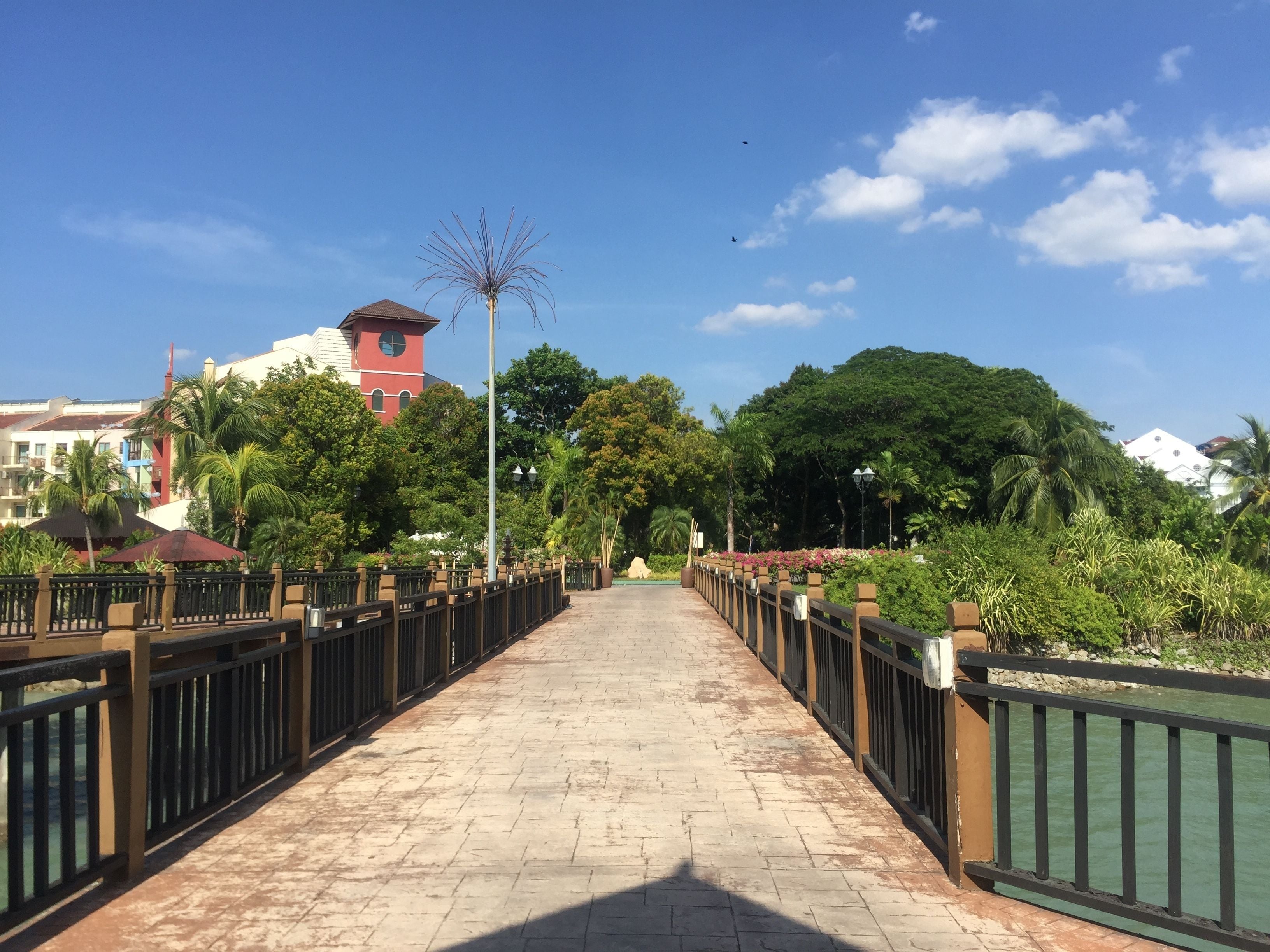 A paved walkway with a metal railing, surrounded by greenery and buildings in the background.
