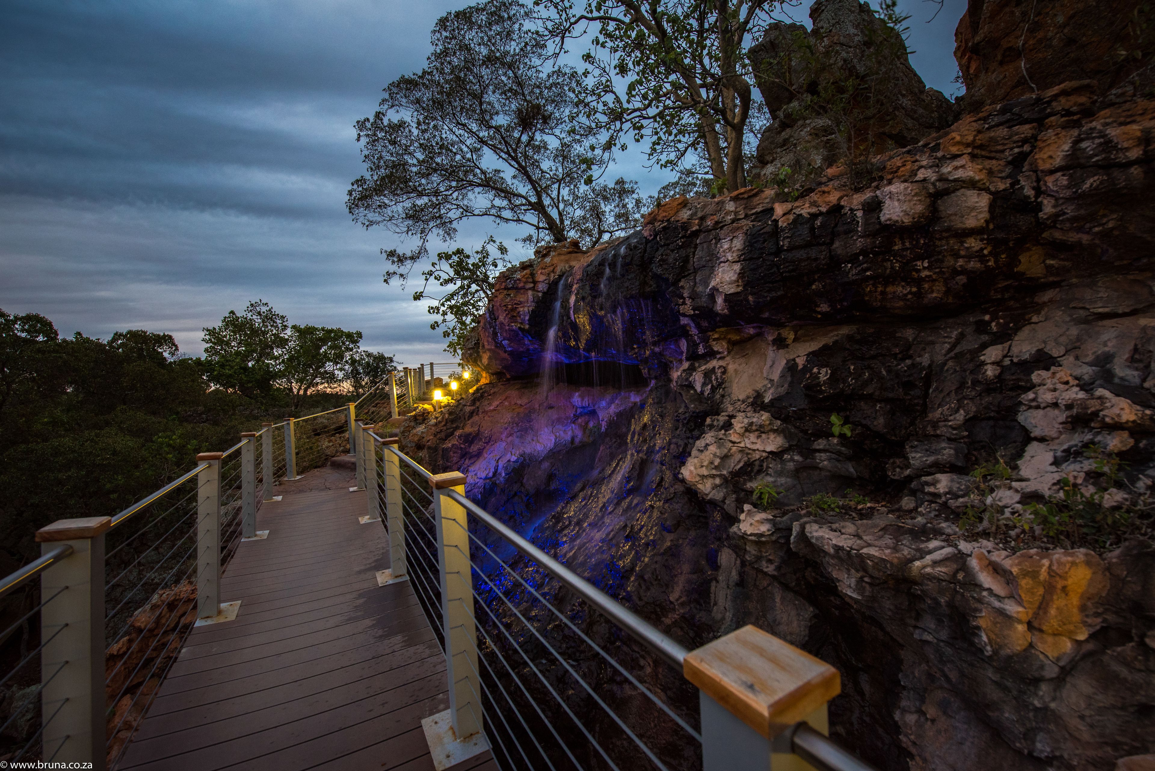 A wooden boardwalk with metal railings leading to a rocky cliff with a waterfall.