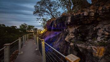 A wooden boardwalk with metal railings leading to a rocky cliff with a waterfall.