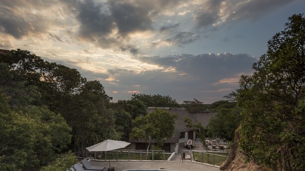 Una piscina con sillas de descanso y sombrillas, rodeada de árboles y un edificio con techo de paja.