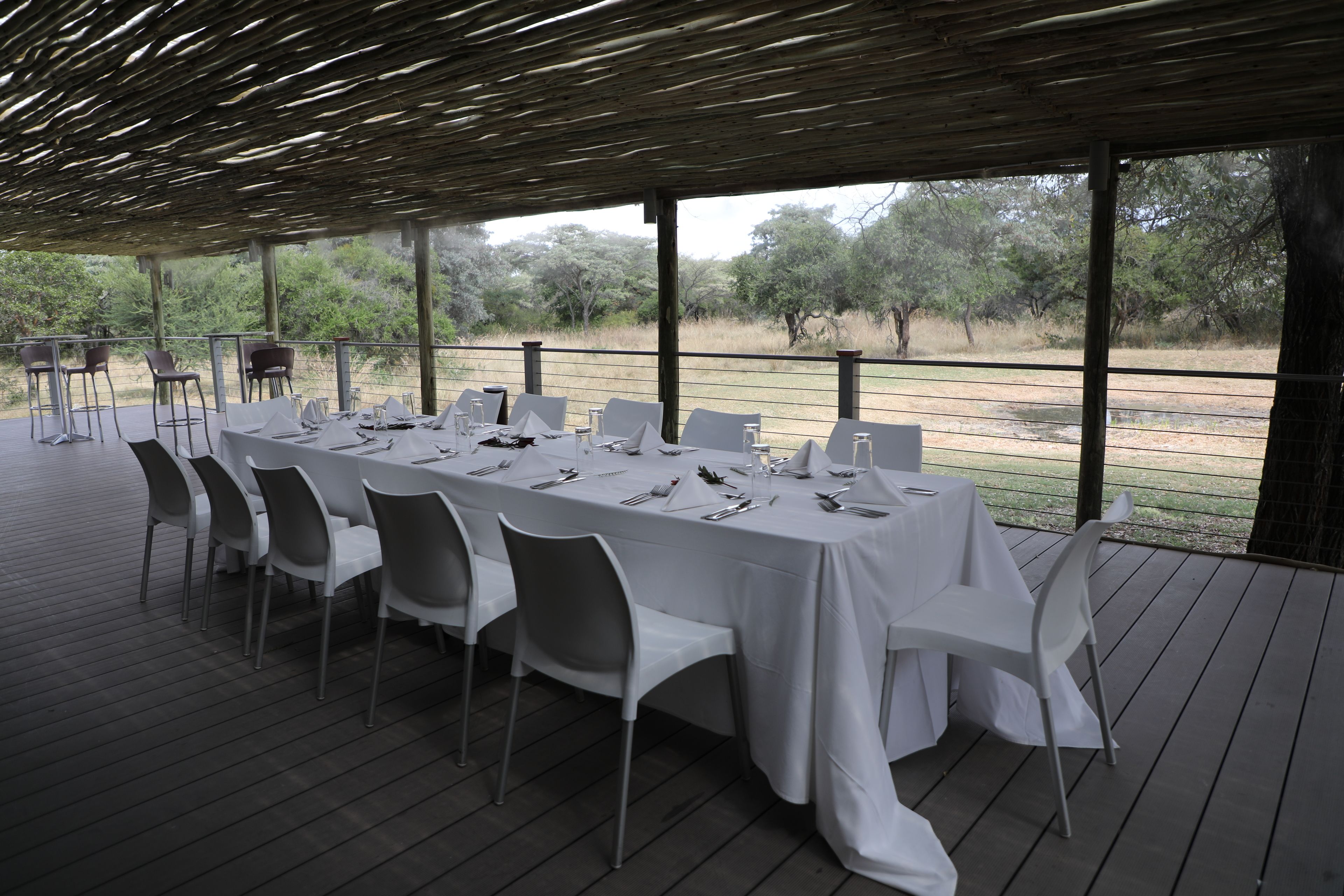 An outdoor dining area with a long table set for six, surrounded by white chairs, under a wooden pergola with a view of the landscape.