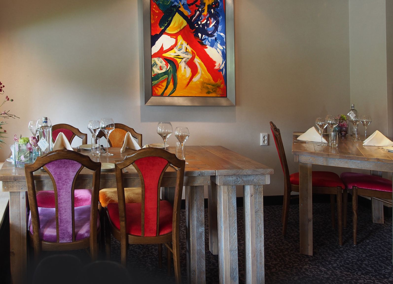 Un espace repas avec des tables et des chaises en bois, des verres à vin et un tableau abstrait aux couleurs vives accroché au mur.