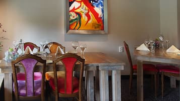 Un espace repas avec des tables et des chaises en bois, des verres à vin et un tableau abstrait aux couleurs vives accroché au mur.