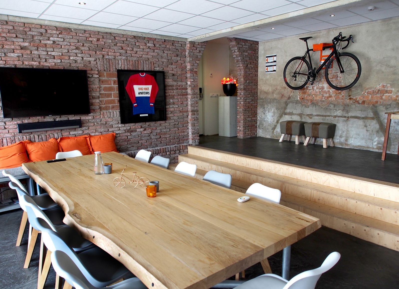 Un salón de conferencias con una larga mesa de madera, sillas blancas, una pared de ladrillo, un televisor montado y una bicicleta sobre un soporte.