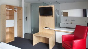 A modern hotel room with a wooden wardrobe, a small wooden table, a red chair, a TV mounted on a wooden panel, and a white desk with a mirror. - Villavelo Twente (Ootmarsum)