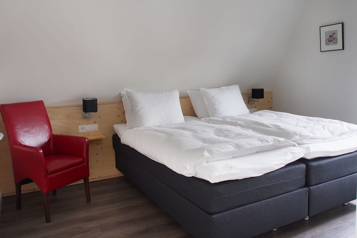 Un dormitorio con cama, una silla roja, una mesita de noche y una lámpara de pared.