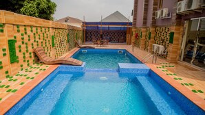 A swimming pool with two lounge chairs, a tiled deck, and a brick wall with green plant decorations. - Maxbe Continental Hotel (Enugu)